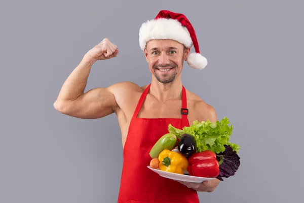 料理エプロンと野菜とサンタの帽子の男 健康的な食事のコンセプト サンタ ビーガン 灰色のスタジオの背景に野菜を持つ若い男 クリスマスや新年のための健康的なビーガン料理 — ストック写真