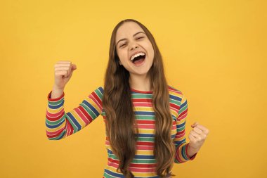 Cocuk kiz seviniyor, evet diyor, mutlu gorunuyor ve zaferi kutluyor, sampiyon jest, yumruk pompasi, sari arkaplanda. Heyecanlı bir yüz, neşeli genç bir kız.