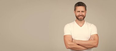 Happy confident man smile keeping arms crossed in white tshirt grey background, guy. Man face portrait, banner with copy space