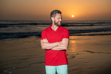 Gün batımında deniz üzerinde sakallı neşeli bir adam. Sahildeki traşsız adam. Sunrise Beach 'teki adam ellerini çaprazladı. Gömlekli sakallı adam. Yaz modası. Yaz tatili. pozitif duygular.