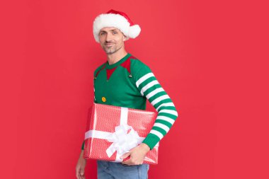 Kırmızı arka planda hediye kutusu olan mutlu Xmas adamı. Mutlu yıllar. Mutlu noel hediyeleri. Elf kostümlü ve Noel Baba şapkalı bir adam..