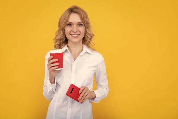 happy blonde woman with coffee cup and smartphone on yellow background.
