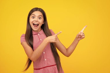 Amazed teen girl. Excited expression, cheerful and glad. Look at advert. Teenager child points aside shows blank copy space for text promo idea presentation, poses against yellow background