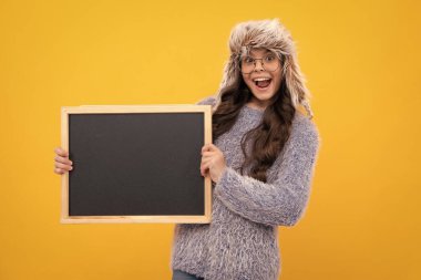 Genç kız, sıcak bir şapka içinde kara tahta tutarak sarı arka planda fotokopi alanı tutuyor. Heyecanlı bir yüz, neşeli genç bir kız.