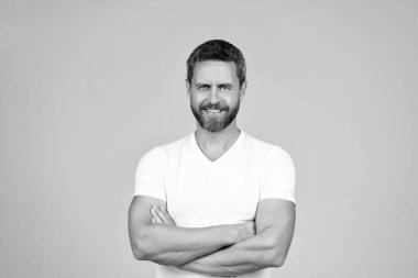 Mutlu, kendine güvenen adam gülümsüyor kollarını beyaz tişörtlü, gri arka planda tutuyor..