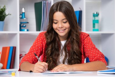 School student writing in exercise book, studying in classroom. Happy schoolgirl