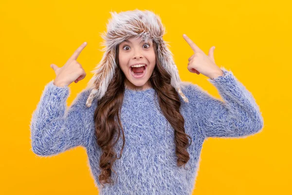黄色の背景にニットを着た10代の女の子 暖かい服を着た子供の肖像画です 肯定的な感情を表現する 冬のファッション 耳フラップの帽子に子供の指がびっくり 女性ファッションモデル — ストック写真