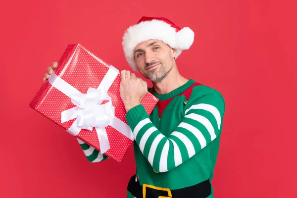 happy curious man in elf costume and santa claus hat. xmas guy with present box on red background. happy new year. merry christmas gift.