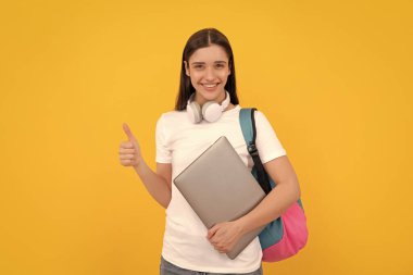 Okul çantalı bir kadın dizüstü bilgisayar tutuyor. İnternetten al. Okula dönüyorum. Başparmağını kaldır. Etkileyici bloglar. Sırt çantası ve kulaklığı olan pozitif bir kız. Modern eğitim. iletişim teknolojisi.