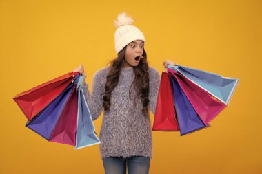 Sarı arka planda alışveriş torbaları olan, kışlık sıcak şapkalı güzel moda ergen kız çocuğu. Alışveriş ve moda alışverişi. Alışveriş çantalı çocuk. Heyecanlı yüz..