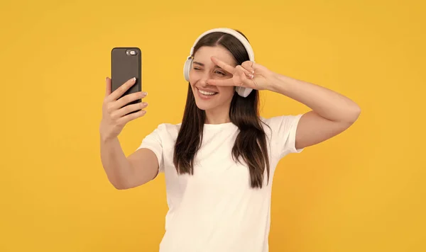 positive lady in white shirt in headphones making selfie on phone with peace gesture, selfie.