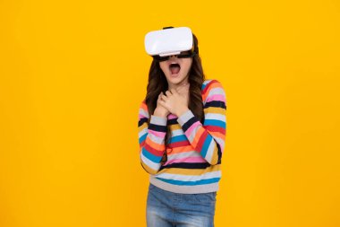 Çocuk için sanal aletler, boş zaman ve eğitim. Genç liseli kız, sarı arka planda izole edilmiş VR gözlükleri kullanıyor, stüdyo fotoğrafı. Şaşırmış yüz, şaşırmış genç kız duyguları.