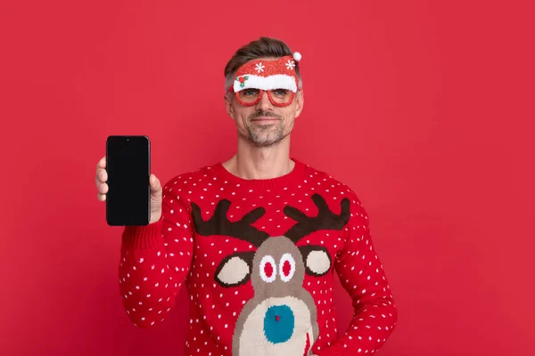 happy man in sweater and party glasses. xmas guy presenting phone screen on red background. happy new year. merry christmas. greeting online.