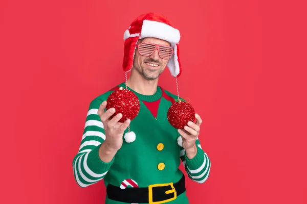 快乐的圣诞节 身穿精灵服装 头戴圣爪帽的快乐男人 戴着派对眼镜 戴着红色背景圣诞装饰球的家伙 新年快乐 — 图库照片