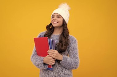 Sırt çantasıyla sarı arka planda not defteri tutan örgü şapkalı ve kalın kazaklı gülümseyen bir kız öğrenci portresi. Mutlu okul kızı, pozitif ve gülümseyen duygular