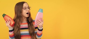 amazed child long hair in glasses choosing between shampoo and conditioner bottle, choice. Banner of child girl hair care, studio poster header with copy space