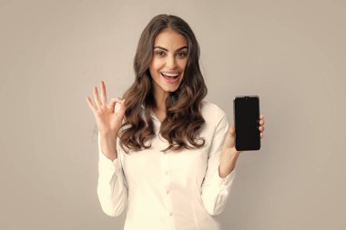 Mutlu genç kadın boş akıllı telefon ekranı gösteriyor. Gülümseyen esmer kadın, gri arka planda izole edilmiş modern teknolojilere bağımlı sosyal ağlarda cep telefonu mesajları kullanıyor.