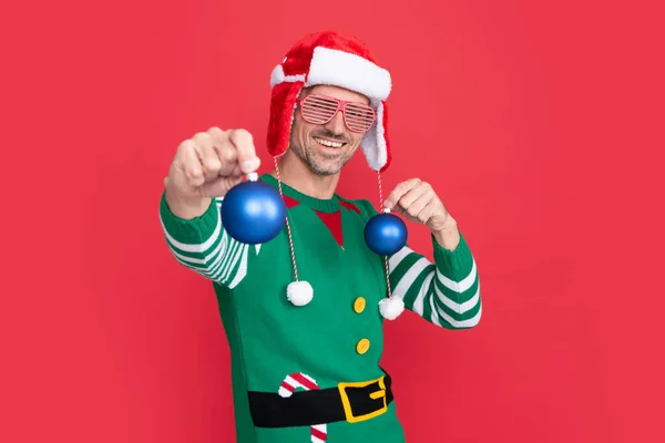 glad man in elf costume and santa claus hat. guy in party glasses hold xmas decoration balls on red background. happy new year. merry christmas.