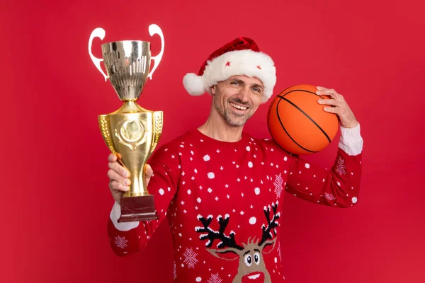Santa trainer hold basketball ball and champion cup. Man in Christmas sweater and Santa hat on red background