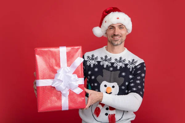 Portrait of Santa man in sweater holding big red present box isolated over red background. Concept of x-mas time celebration holidays, happiness, emotions