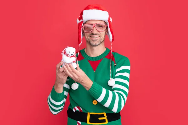 happy man in elf costume and santa claus hat. guy in party glasses hold xmas decoration snowman on red background. happy new year. merry christmas.