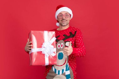 Kırmızı arka planda hediye kutusu olan sürpriz xmas adamı. Mutlu yıllar. Mutlu noel hediyeleri. Başparmağını kaldır. Kışın süveter ve Noel Baba şapkalı bir adam..