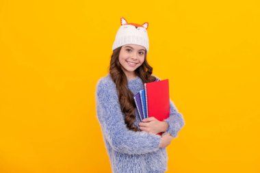 Çalışma kitabını tutan çocuğun portresi. Olumlu duygularını ifade et. Kış modası. Şapkalı, kıvırcık saçlı, gülümseyen çocuk. Okula dönüyorum. Sarı arka planda örgü elbiseli genç kız..