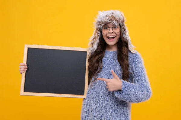 School sales board. Teen schoolgirl in warm winter hat hold blackboard. Back to school. Excited face, cheerful emotions of teenager girl