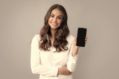 Mutlu genç kadın boş akıllı telefon ekranı gösteriyor. Genç bir kadın cep telefonundan mesaj okuyor ve gülümsüyor. Akıllı telefonlu mutlu iş kadını, gri arka planda duruyor.