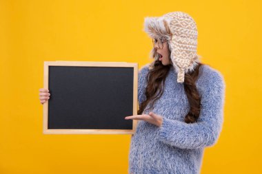 Sarı arka planda izole edilmiş, kalın bir şapka ve kazak giyen genç bir kız çocuğu. Kış okulu satışı, fotokopi alanı, maket. Şaşırmış yüz, şaşırmış duygular