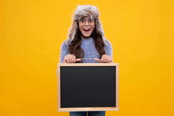 Teenager girl hold in warm winter hat hold blackboard chalkboard with copy space on yellow background. Excited face, cheerful emotions of teenager girl