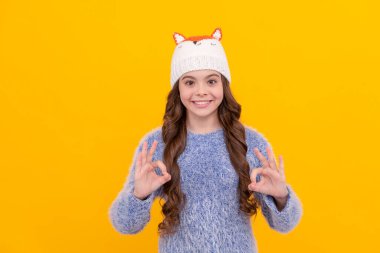 Kış modası. Tamam. Şapkalı, kıvırcık saçlı mutlu çocuk. Kadın manken. Sarı arka planda örgü giyen genç bir kız. Kalın giysiler giyen çocuk portresi. pozitif duygularını ifade et.