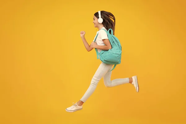 School girl, teenager student in headphones on yellow isolated studio background. Music school concept. Excited teenager, school leisure. Jump and run, jumping child