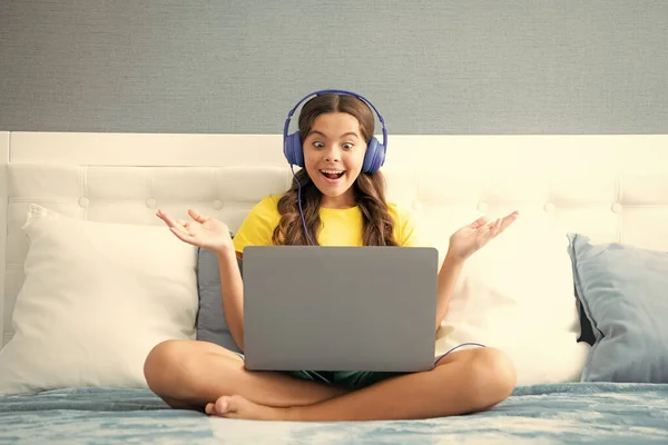 Teenager girl in headphones relax on bed at home using laptop. Child in earphones browse internet on computer. Expressive emotional excited teen girl