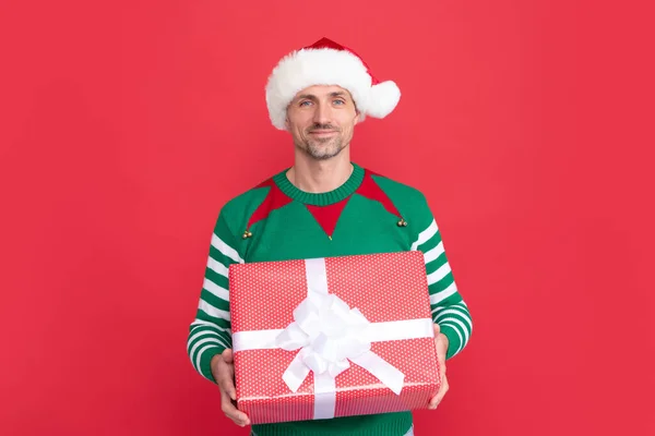 smiling man in elf costume and santa claus hat. xmas guy with present box on red background. happy new year. merry christmas gift.