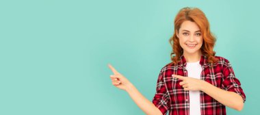 Mavi arka planda kareli tişörtlü mutlu kızıl kadın işaret ediyor. İzole edilmiş yüz portresi, kopyalama alanı oluşturulmuş pankart