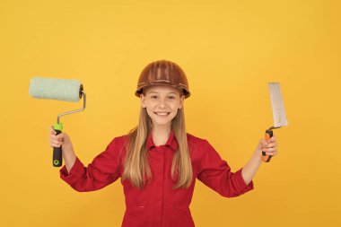 cheerful teen child in builder hard hat with paint roller and spatula on yellow wall.