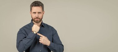 Unshaven businessman in casual shirt point finger at wrist watch grey background, business hour. Man face portrait, banner with copy space