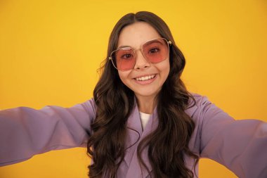 Güneş gözlüklü genç kız sarı arka planda poz vererek selfie çekiyor. Moda ergeni, sıradan görünüm.