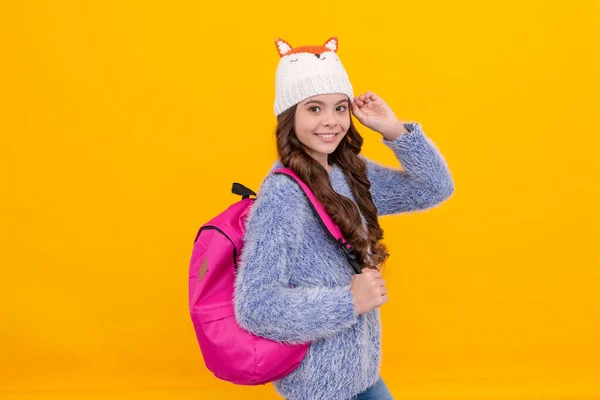 express positive emotion. winter fashion. positive kid with curly hair in hat. back to school. teen girl in knitwear on yellow background. portrait of child wearing warm clothes carry backpack.