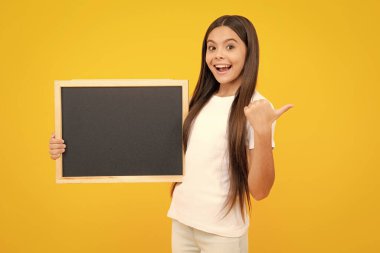 Teenager child holding blank chalkboard for message Isolated on a yellow background. Empty text blackboard, copy space mock up. Happy teenager portrait. Smiling girl