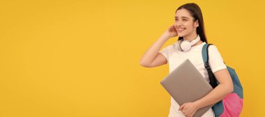 Sırt çantalı neşeli genç kız sarı arka planda kulaklıklarla dizüstü bilgisayarı tutuyor, e-öğreniyor. İzole edilmiş yüz portresi, kopyalama alanı olan pankart