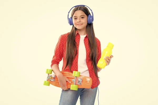Teenagers lifestyle, casual youth culture. Teen girl with skateboard water bottle and headphones over isolated studio background. Cool modern teenager in stylish clothes