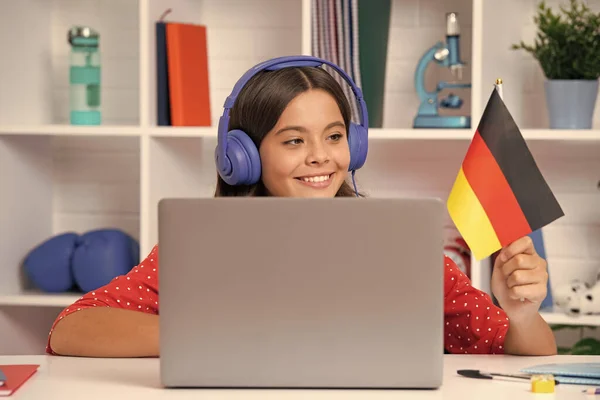 A young school girl student sitting at the table, using laptop when studying. Happy girl hold german flag, positive and smiling emotions