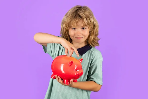Saving money concept. Portrait of a little boy putting money on a moneybox. Child saving money in a piggybank on studio background