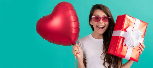 amazed child in eyeglasses hold gift box and valentines day party heart balloon, love day. Teenager girl with birthday gift, horizontal poster. Banner header with copy space