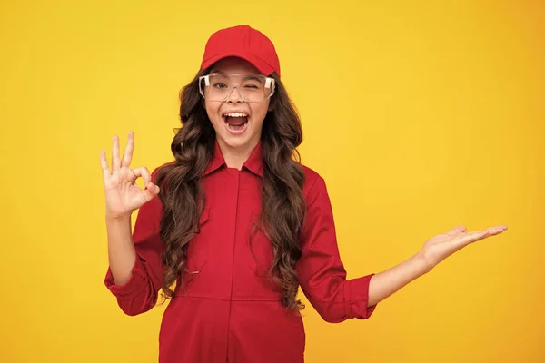 Excited teenager girl. Worker teenager child wearing overalls red, cap and protect glasses. Studio shot portrait isolated on yellow background. Pointing and showing concept
