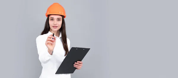 Young woman in hard hat helmet with clipboard isolated on grey background. Architect woman wear helmet and shirt. Woman isolated face portrait, banner with mock up copy space