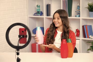 Çocuk video kaydedicisi telefonda video blogu yapıyor. Web günlüğü ve vlog. Genç kız blogger etkileyicisi sosyal medya için saç kremi ya da şampuan şişesi tutuyor. Mutlu kız yüzü, pozitif ve gülümsüyor.