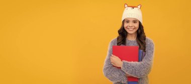 Kış okulu. Sarı arka plandaki süveter ve şapka tutma kitabı. Öğrenci portresi, stüdyo afişi. Okul çocuğu yüzü, boşluklar